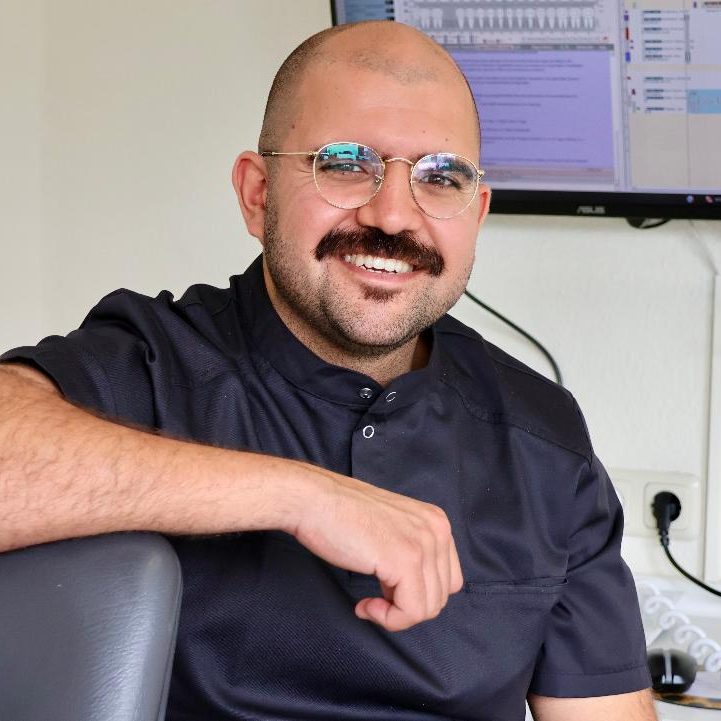 @FachzahnarztKoeln Instagram Lächelnder Mann mit Brille und Schnurrbart, sitzt in einem modernen Zahnarztbehandlungsraum.