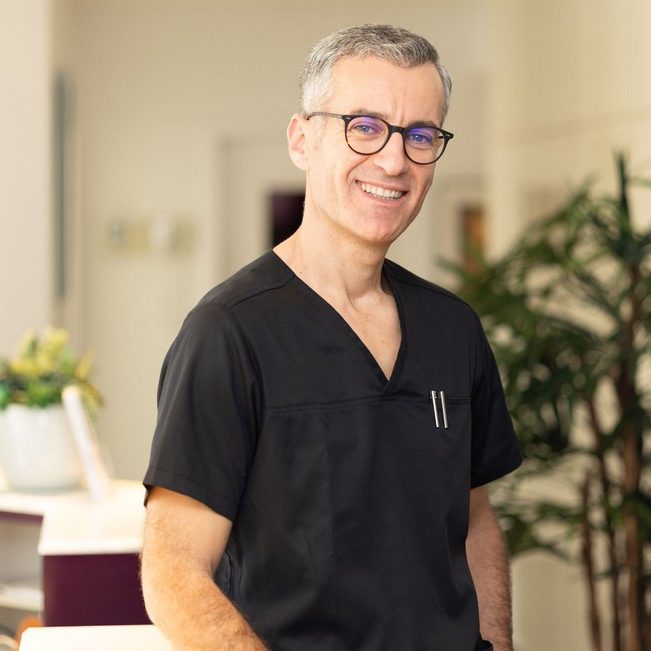 @FachzahnarztKoeln Instagram Lächelnder Mann mit Brille und Schnurrbart, sitzt in einem modernen Zahnarztbehandlungsraum.