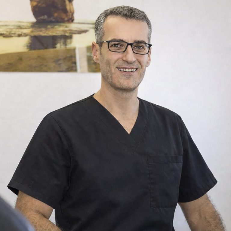 @FachzahnarztKoeln Instagram Lächelnder Mann mit Brille und Schnurrbart, sitzt in einem modernen Zahnarztbehandlungsraum.